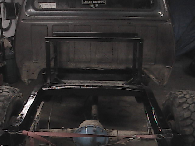Rear view of a truck frame with a custom metal rack welded onto it. Large tires are visible