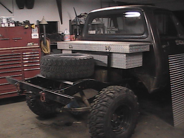 Truck chassis with large spare tire, toolbox, and custom rear bed frame. Dark color, appears to be in a garage