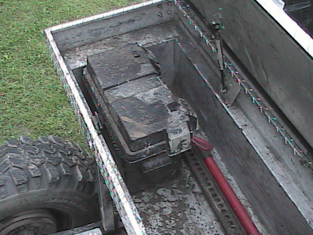 Truck bed with battery box and tire. The battery box is dirty and contains a large battery. A red tool is visible inside the box. The tire is large and off-road style