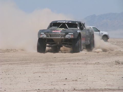 2008 Ford Bronco - A black off-road truck races through the desert, kicking up a cloud of dust as a white SUV follows behind.