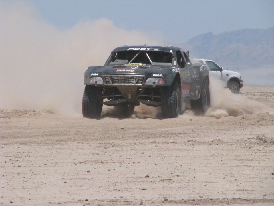 Off-road race truck kicking up dust, another white SUV behind it on a dirt track