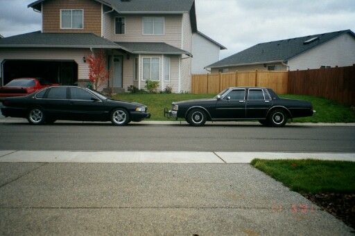 Two dark full-size sedans parked in side view on a suburban street by a tan house