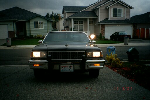 Dark full-size Chevrolet sedan, front view, headlights on in driveway at dusk.