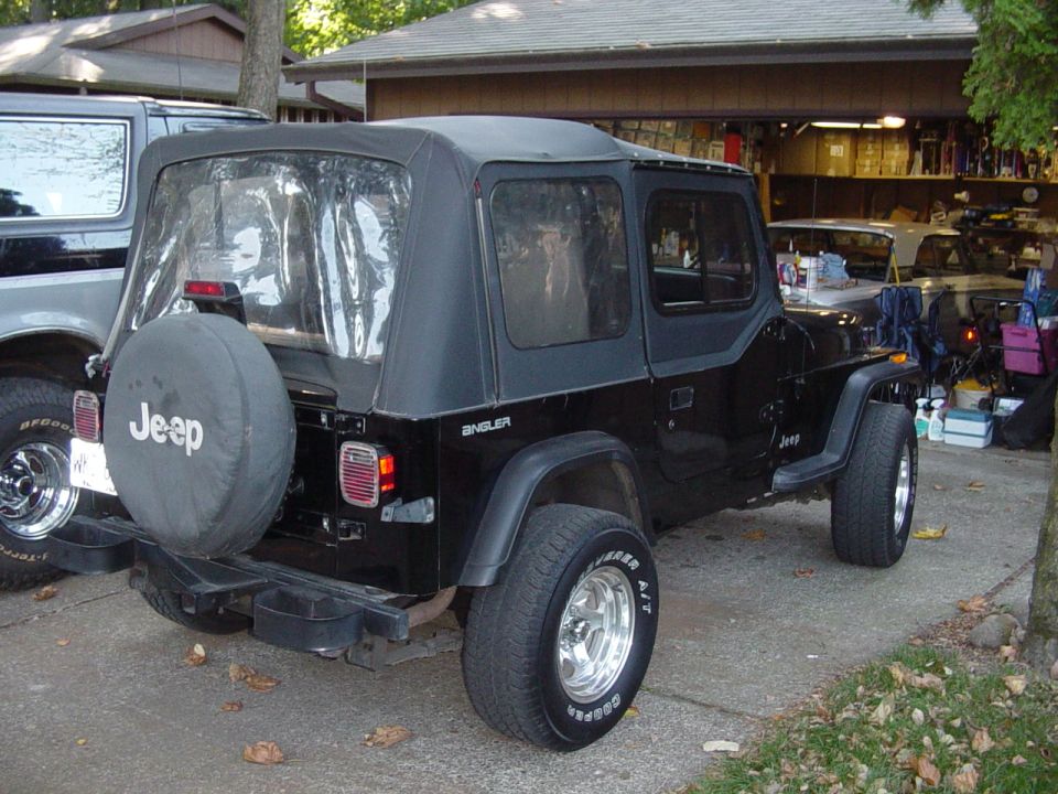 Black Jeep Wrangler with soft top and Jeep spare tire cover, rear three-quarter view in a driveway