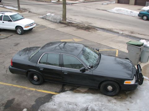 2000 Ford Crown Victoria - A black 2000 Ford Crown Victoria police car is parked next to a white SUV in a lot with snow on the ground.