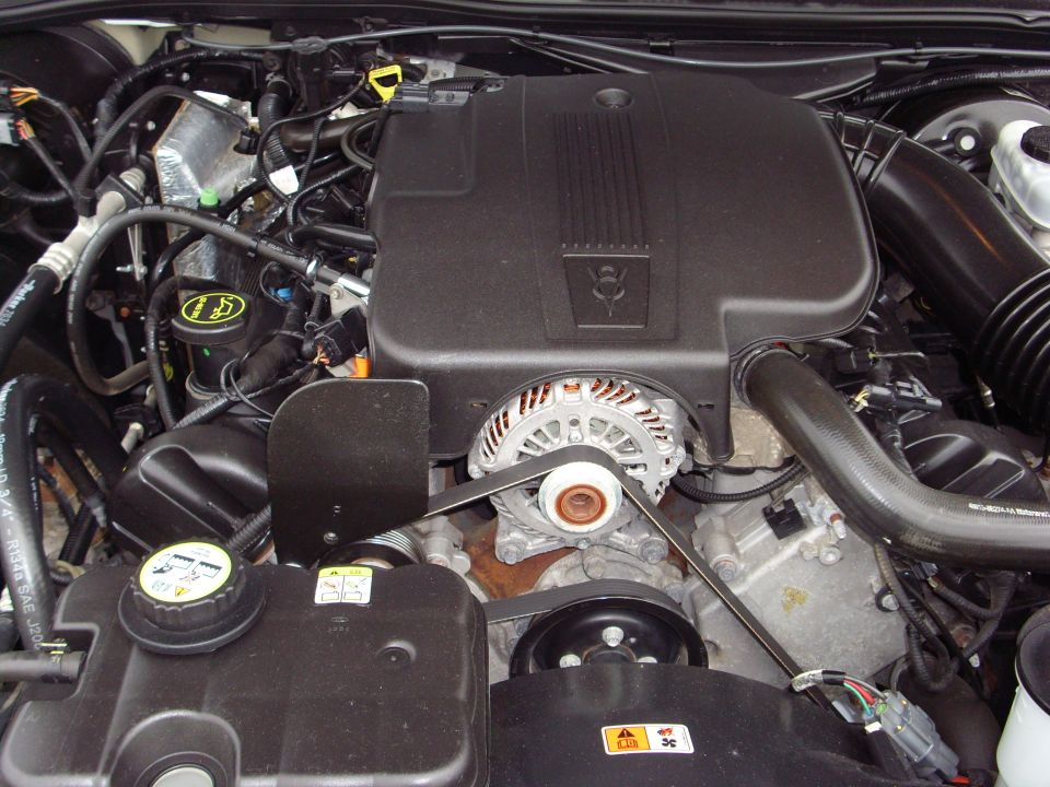 Engine bay with black plastic covers, alternator, belts, and hoses. A SuperMotors.net watermark is visible