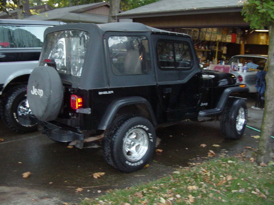 Black Jeep Wrangler soft top from rear quarter, Jeep spare tire cover, parked on wet leaf-covered driveway