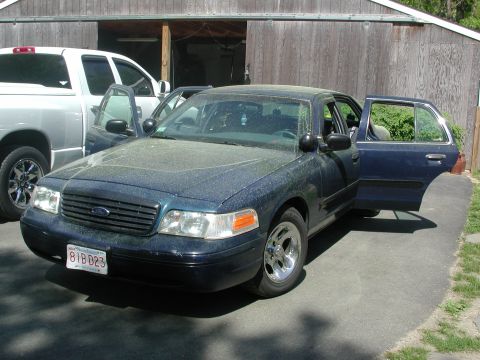 1999 Ford Crown Victoria - A dark blue Ford Crown Victoria is parked in front of a wooden garage with its doors open.