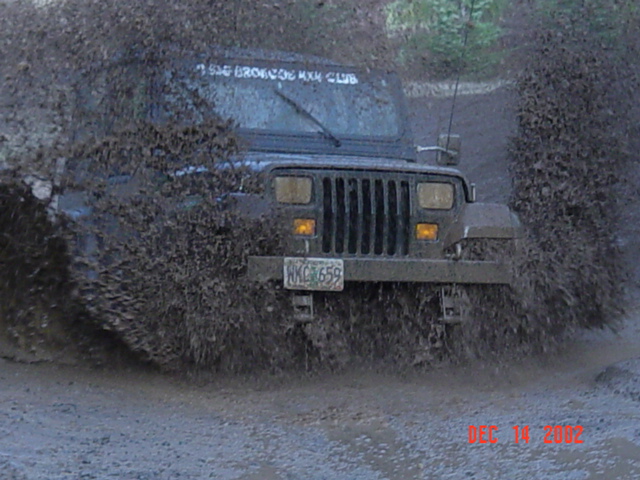 Dark Jeep Wrangler blasting through a deep mud puddle, water spraying high on both sides on a trail