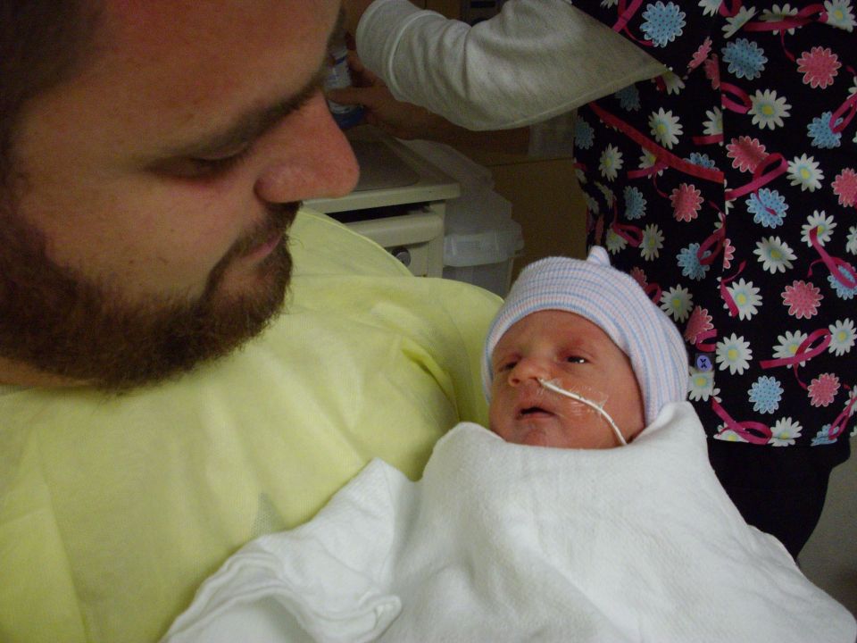 Newborn baby wrapped in a white blanket, wearing a blue cap, with a feeding tube, held by a man with a beard
