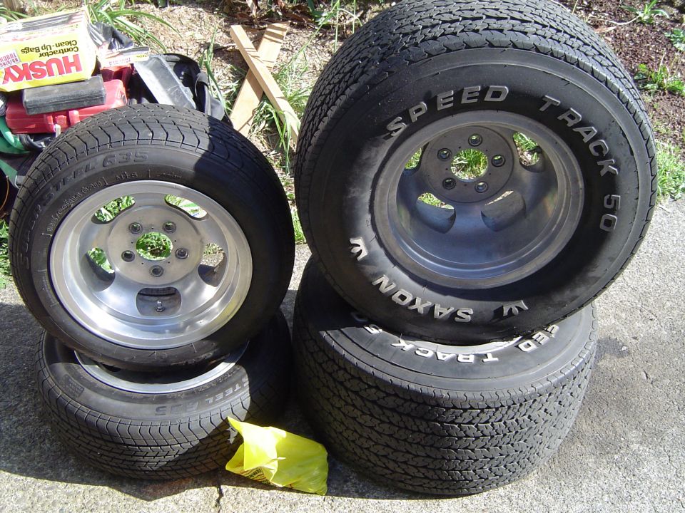 Four used 5-lug alloy wheels on white-letter tires stacked in pairs on a concrete driveway