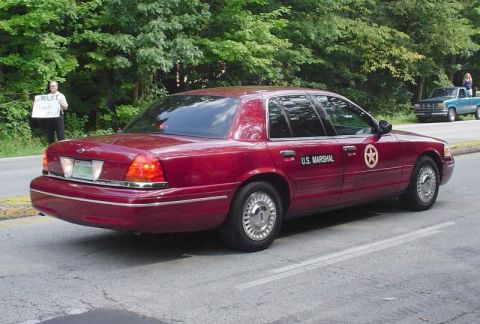1991 Ford Crown Victoria - Maroon Ford Crown Victoria marked as U.S. Marshal, with a person holding a sign in the background.