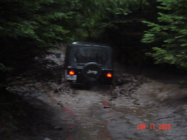 Dark Jeep Wrangler from behind crossing a flooded muddy trail at dusk, brake lights glowing