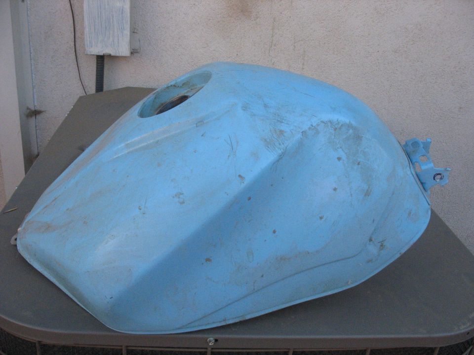 Light blue motorcycle fuel tank with scuffs and scratches, viewed from the side