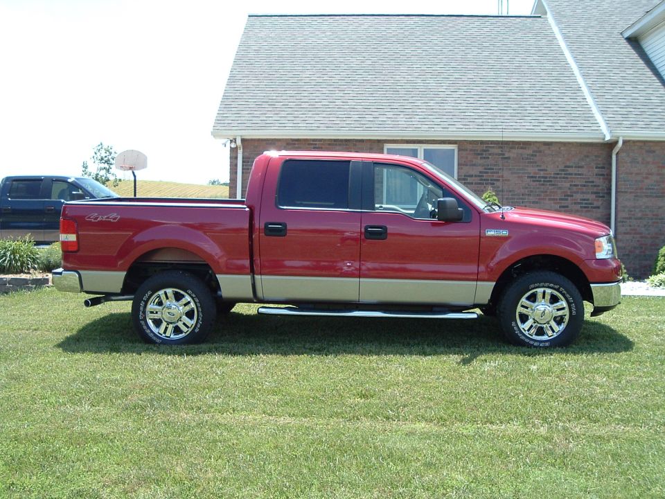 Red four-door pickup truck parked on grass in front of a house. Another dark-colored truck is visible in the background