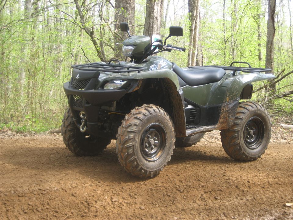 Green Suzuki utility ATV with winch and brush guard on a dirt forest trail