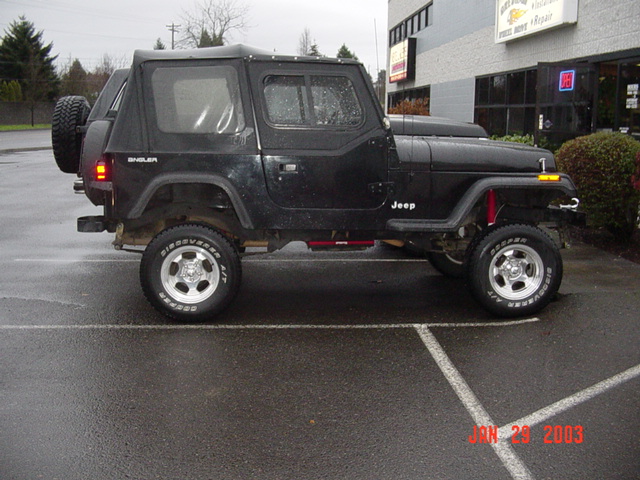Driver side of lifted black Jeep Wrangler with soft top and chrome wheels on wet lot