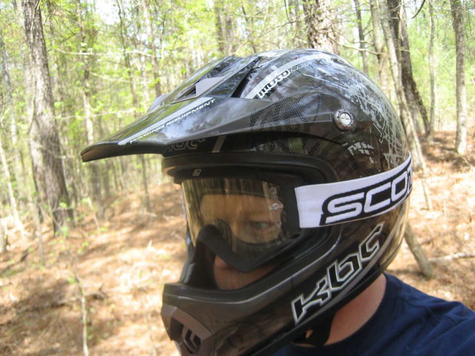 Black dirt bike helmet with white Scott strap and reflective visor in a forest.