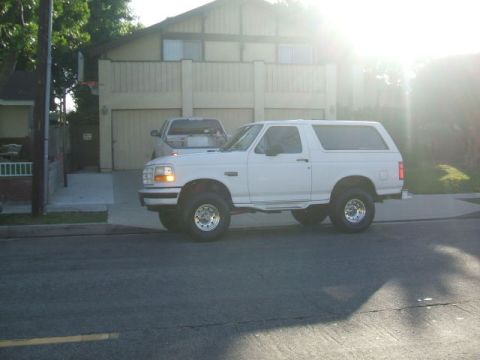 1988 Ford Bronco - A white 1988 Ford Bronco is parked on the street in front of a two-story house with a garage. The sun is shining brightly, creating a lens…