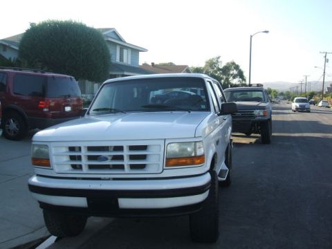 1988 Ford Bronco - A white Ford Bronco is parked on a residential street in front of a maroon SUV. The Bronco is facing the camera with its front grille and h…
