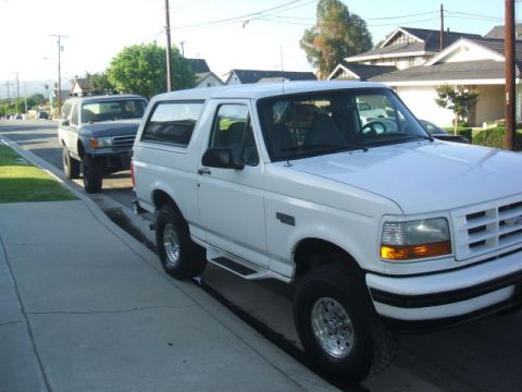 1988 Ford Bronco - A white Ford Bronco is parked on the street next to a dark gray Ford Bronco. The white Bronco is in the foreground, showing its front quart…