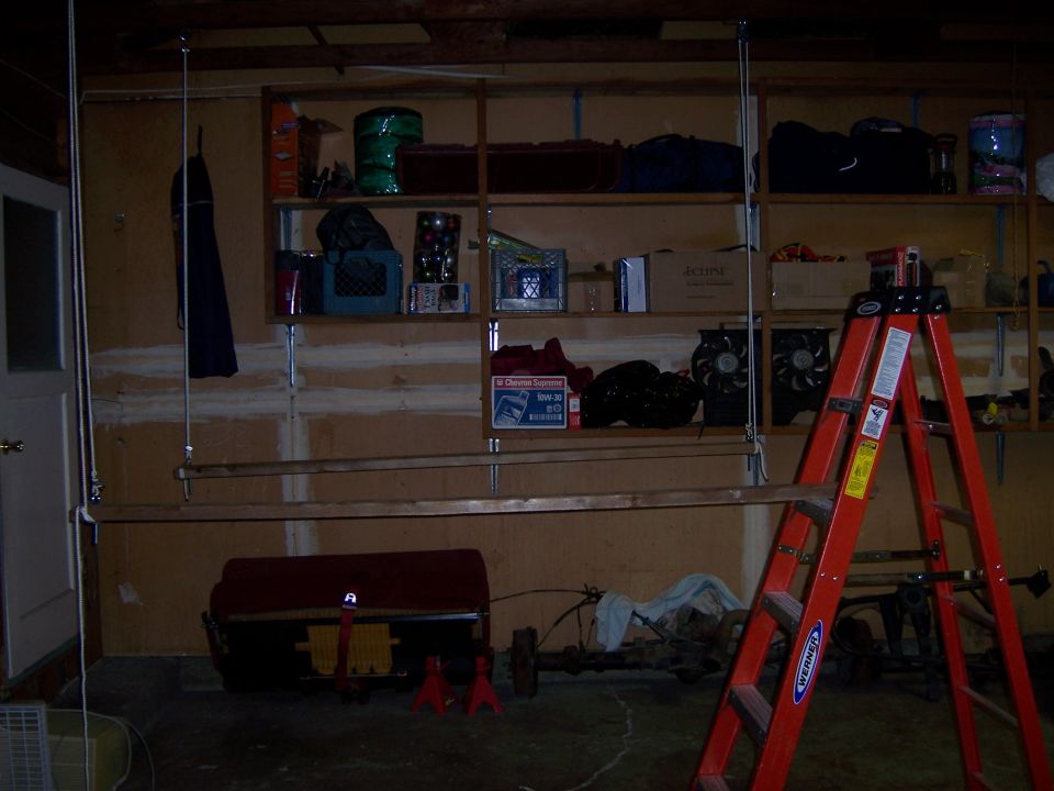 Garage interior with shelves holding storage bins, a red toolbox, vehicle parts on the floor, and a red Werner ladder