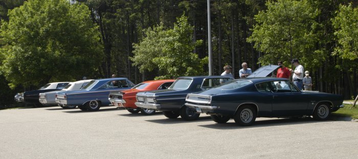 Several classic cars parked in a line, with trees and people in the background