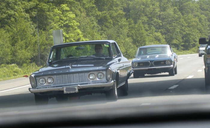 Dark blue 1962 sedan driving on a highway, front view. Grille has horizontal bars and dual headlights