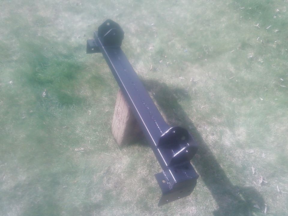 Black metal frame part with mounting brackets, resting on a wooden block on grass