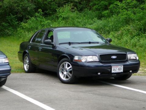 1997 Mercury Grand Marquis - A dark blue Ford Crown Victoria is parked on asphalt with a wooded area in the background.