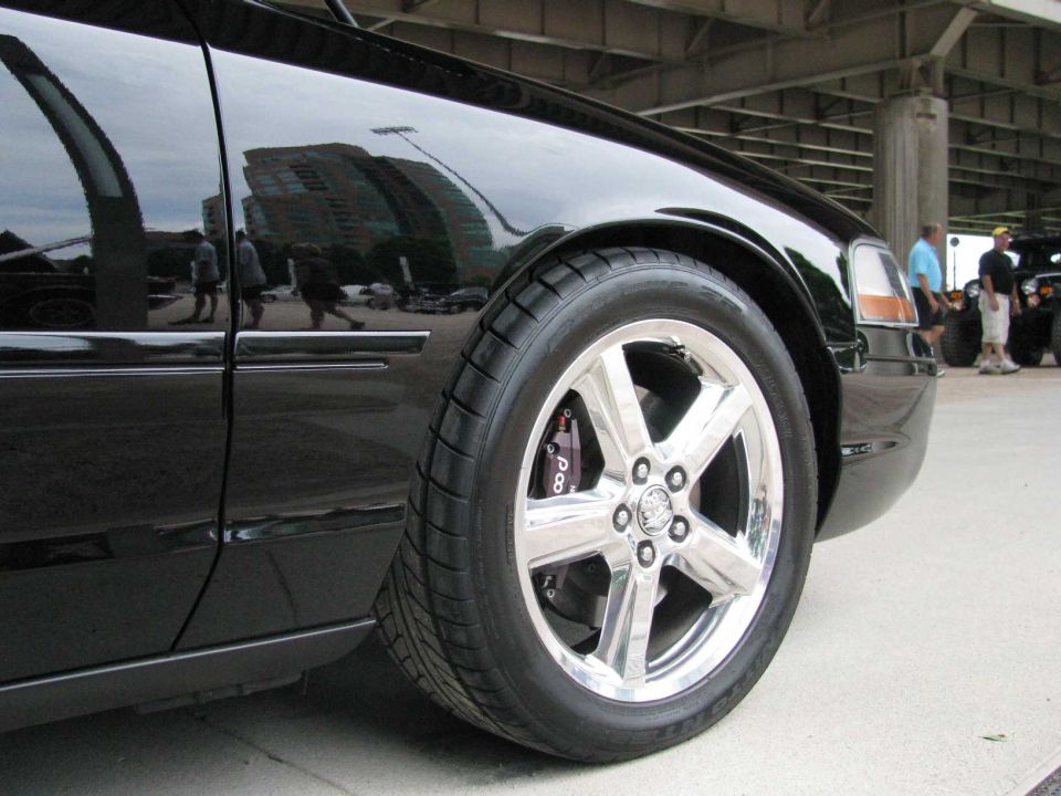 Close-up of a black sedan's front wheel and fender. The wheel is chrome with 5 spokes. The brake caliper is visible and says 'P8'