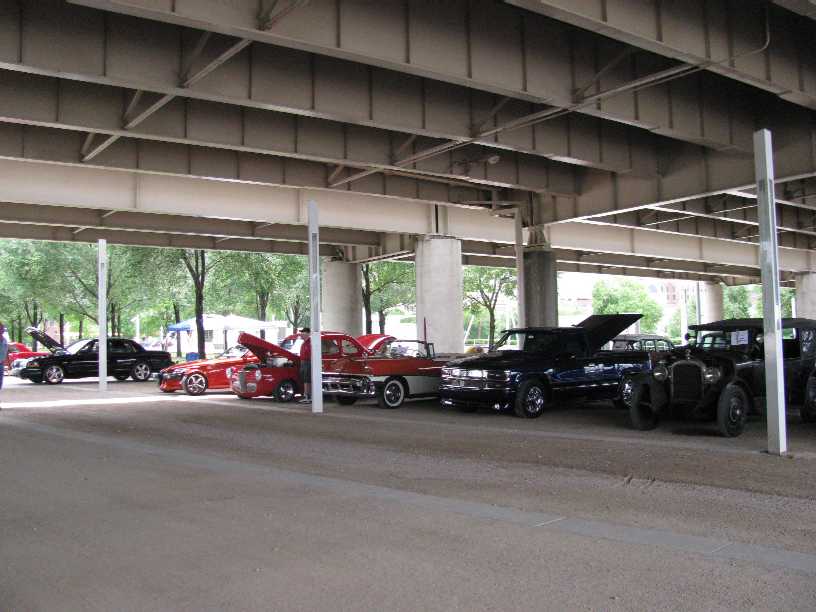 Several cars, including a black sedan with hood up and older models, parked under a concrete overpass