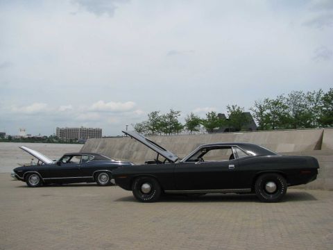 2003 Mercury Marauder - Two black muscle cars with hoods up, parked outdoors near a waterfront. One is a 1970 Plymouth Barracuda.