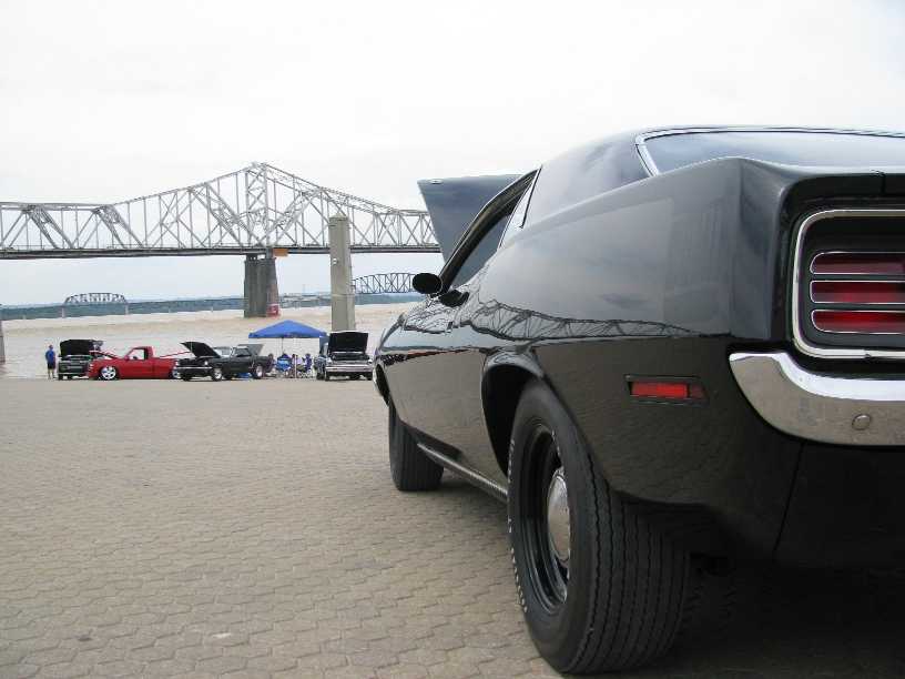 Black muscle car, rear view, parked on paved area near river and bridge. Other cars visible in background