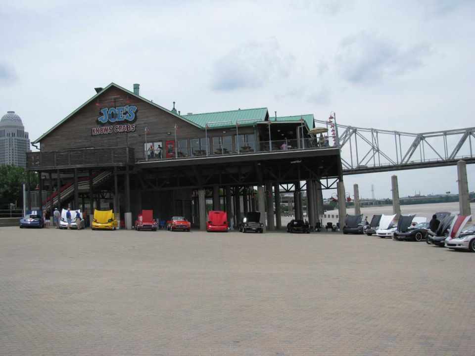 Classic cars parked in front of a waterfront restaurant with a bridge in the background.