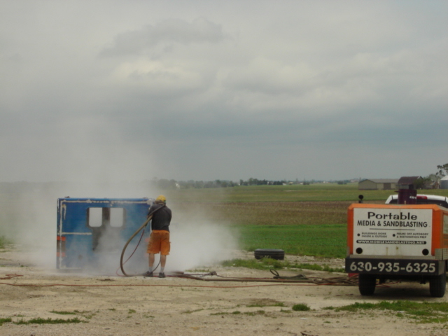 Photo of a worker sandblasting a structure outdoors with a trailer rig