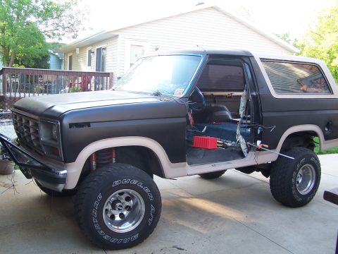 1986 Ford Bronco - A matte black 1986 Ford Bronco with tan trim is parked outside a house with its driver's side door open, revealing the interior and some wi…