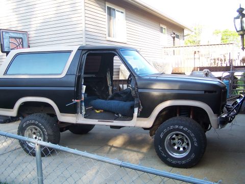 1986 Ford Bronco - The side view of a black 1986 Ford Bronco with a tan top and fender flares is parked outside a house. The driver's side door is open, revea…
