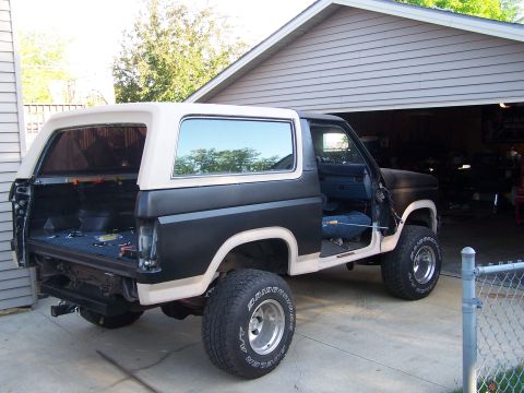 1986 Ford Bronco - A black and tan Ford Bronco is parked in a driveway in front of an open garage. The rear of the vehicle is open, revealing the cargo area,…