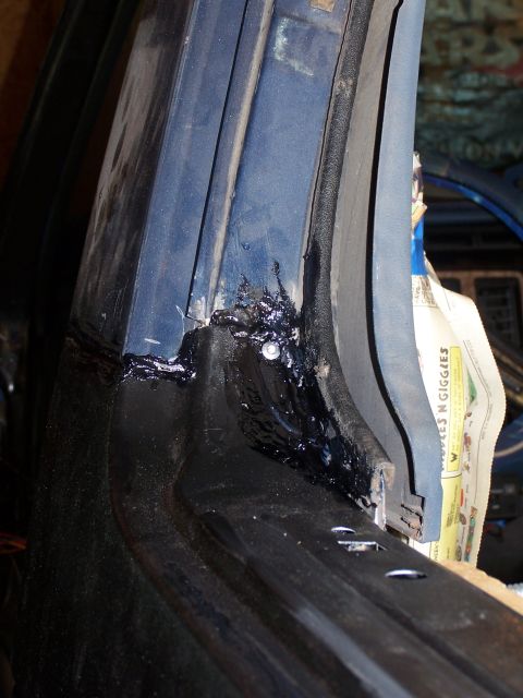 1986 Ford Bronco - A close-up view of a dark blue car door frame with black sealant applied around the edges and a screw visible. A piece of newspaper is tuck…