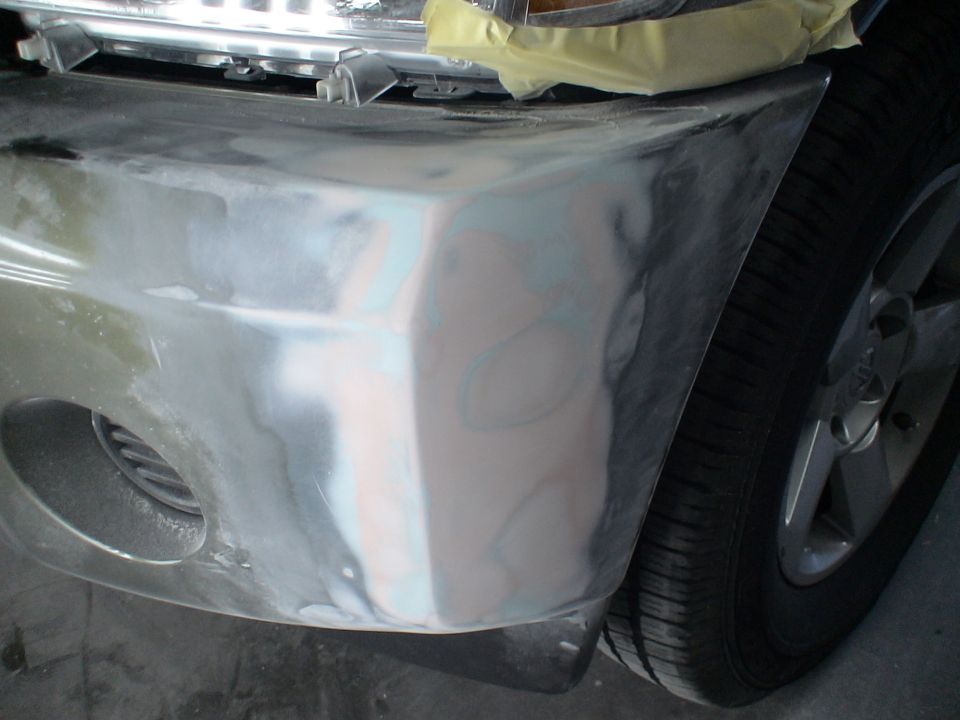 Close-up of a vehicle's front bumper with primer and body filler, next to a tire.