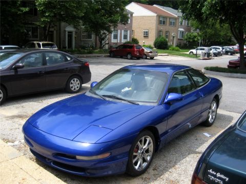 1993 Ford Probe - A bright blue 1993 Ford Probe is parked in a gravel lot in front of apartment buildings. The car is viewed from a low angle, showing its fr…