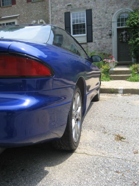 1993 Ford Probe - The rear quarter panel and taillight of a blue 1993 Ford Probe are visible from a low side angle, parked on a gravel driveway in front of a…