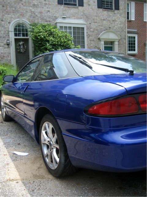1993 Ford Probe - A blue 1993 Ford Probe is parked on a gravel driveway in front of a brick building. The rear passenger side of the coupe is visible, showin…