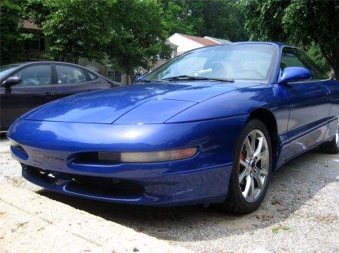 1993 Ford Probe - A low-angle, front 3/4 view of a shiny blue 1993 Ford Probe is parked on a gravel surface with a dark sedan visible behind it. The car feat…