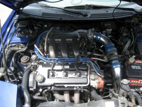 1993 Ford Probe - The engine bay of a blue 1993 Ford Probe is shown with the hood open. Visible modifications include a chrome intake pipe with blue couplers…