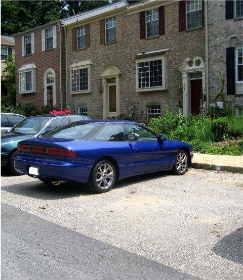 1993 Ford Probe - A blue 1993 Ford Probe is parked in a gravel lot in front of a brick townhouse. The car is viewed from the rear passenger side.