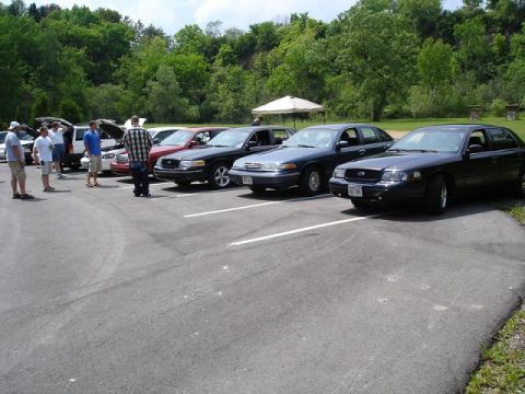 1997 Mercury Grand Marquis - A line of sedans are parked in a lot with trees and a small canopy in the background. Several people are standing near the vehicles.