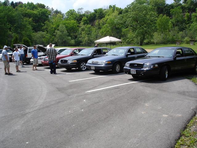 Several sedans parked in a lot with people standing around them.