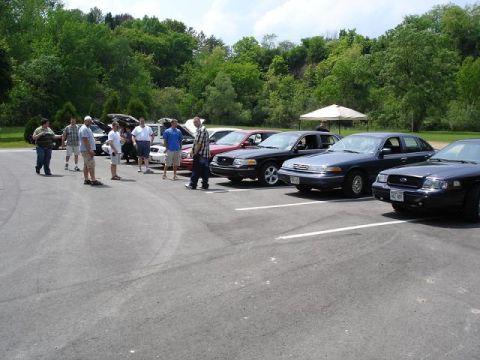1997 Mercury Grand Marquis - A group of people stand around several parked sedans in a parking lot with a wooded hillside in the background.